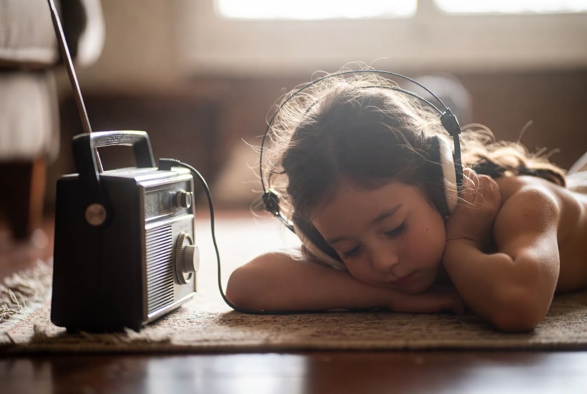 Niña pequeña tumbada en el suelo escuchando una radio antigua con auriculares, en un ambiente cálido y tranquilo.